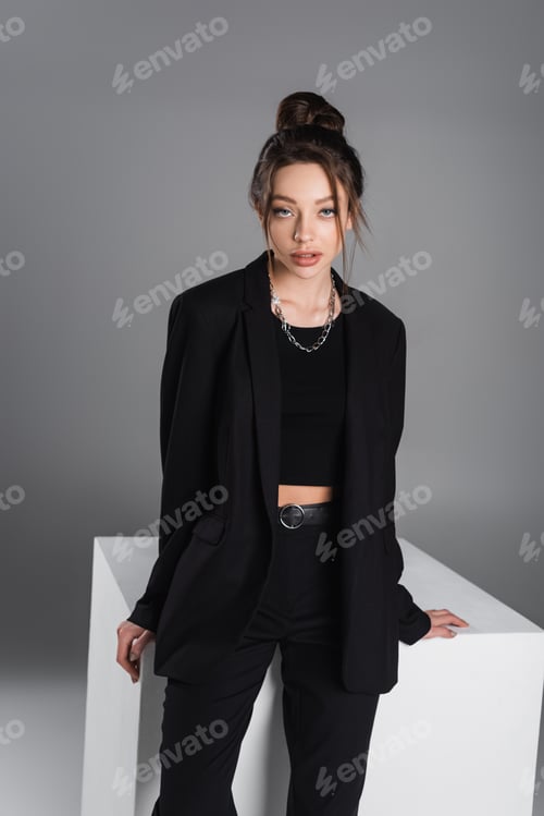 Preview: brunette woman in black suit and silver necklace looking at camera near white cube on grey