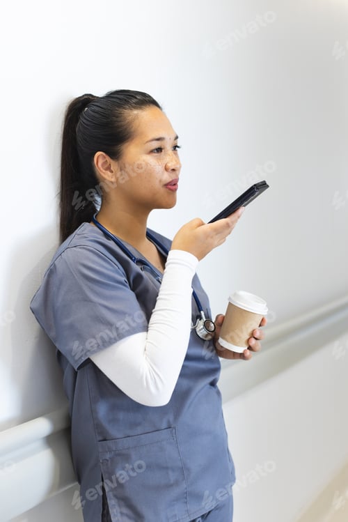 Preview: Biracial female doctor talking on smartphone, holding takeaway coffee in hospital corridor
