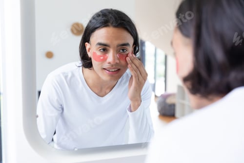 Preview: Happy biracial man looking in mirror, applying eye patches in bathroom