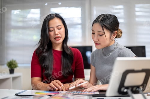 Preview: Asian woman explaining color chart to friend or coworker aside tablet on table in home office studio