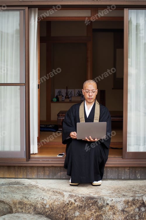 Preview: Buddhist monk sitting outside temple looking at laptop computer.