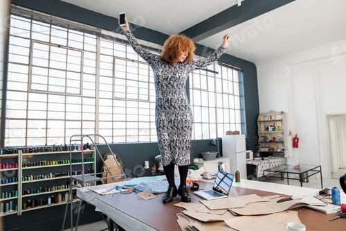 Preview: Mature female fashion designer dancing to smartphone music on workshop table