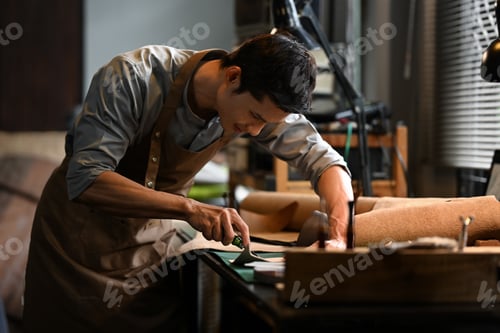 Preview: Small business craftsman in apron carefully cutting piece of leather in his workshop