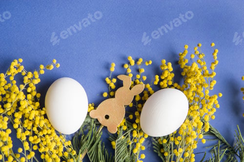 Preview: Easter background, white eggs and a wooden hare on a blue background, decorated with Mimosa flowers,