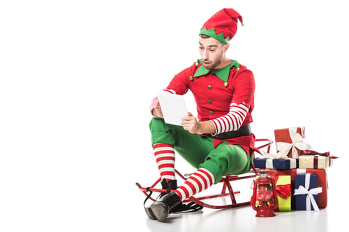 Preview: surprised man in christmas elf costume sitting on sleigh near pile of presents and looking at