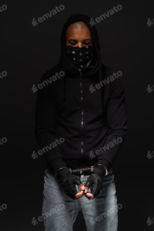 Preview: African american bandit with mask on face and handcuffs standing isolated on black