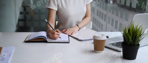 Preview: Cropped shot of businesswoman sitting in modern workplace and writing business plan on notebook.