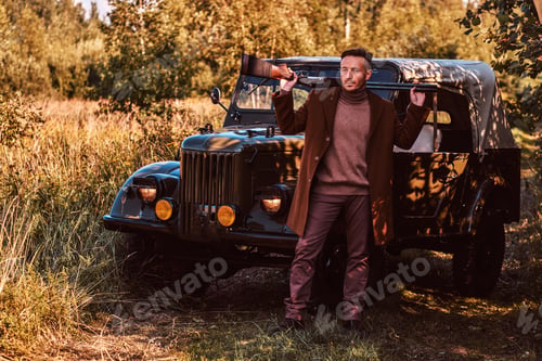 Preview: Old fashioned hunter standing together with his beagle dog near a retro military car in a forest.
