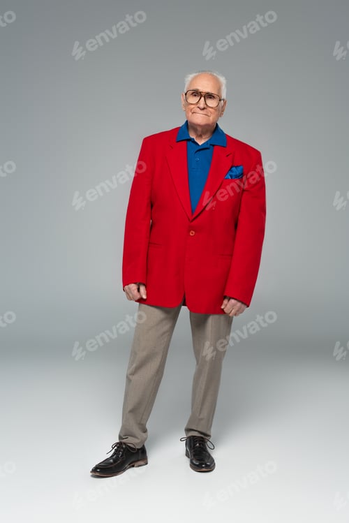 Preview: full length view of serious elderly man in glasses, red blazer and blue shirt standing on grey