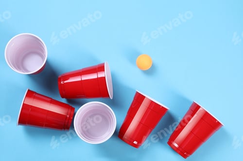 Preview: Plastic cups and ball on light blue background, flat lay. Beer pong game