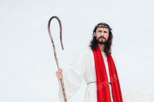 Preview: low angle view of Jesus in robe, red sash and crown of thorns standing with wooden staff in desert
