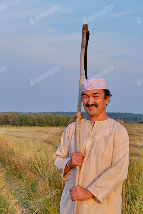 Preview: Smiling senior muslim man with hand-scythe in hayfield.