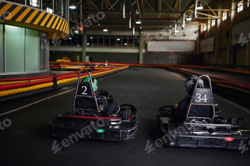Preview: two karting drivers in helmets and sportswear driving go kart on indoor circuit, competitors
