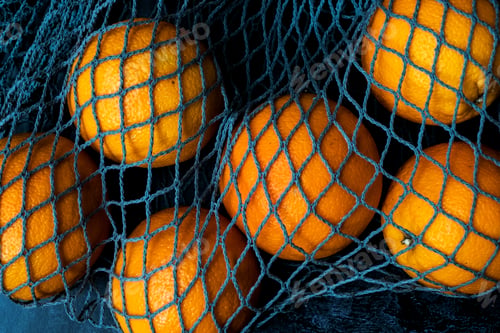 Preview: High angle close up of oranges in grey net bag on black background.