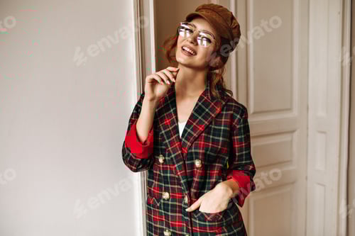 Preview: Charming woman in eyeglasses poses in white apartment. Modern girl with wavy hair in brown cap and