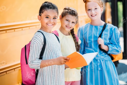 Preview: adorable little schoolgirls with notebook looking at camera in front of school bus