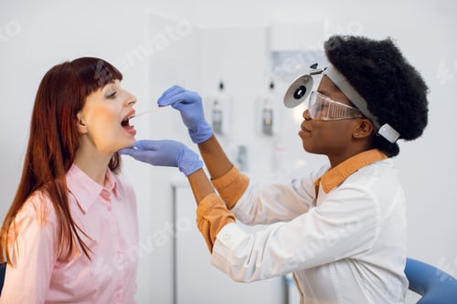 Preview: Young afro-american female ent doctor examining young european woman larynx in modern office. Black