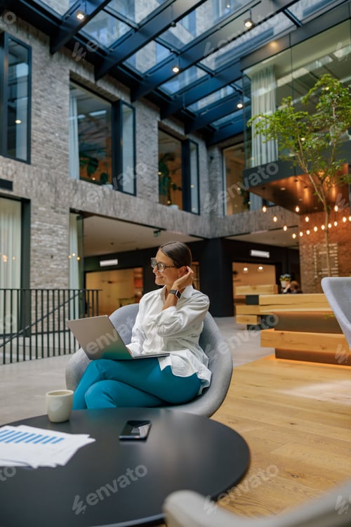 Preview: Smiling stylish business woman working on laptop sitting on modern office background