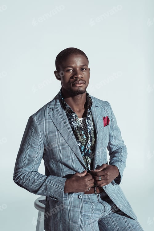 Preview: Stylish african american man in suit, leaning on a bar stool and looking at camera, isolated on grey