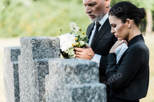 Preview: selective focus of man with grey hair holding flowers near tombstones and woman