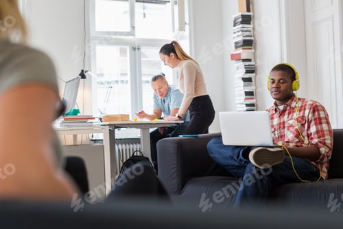 Preview: Worker using laptop on sofa and listening music, colleagues working in background