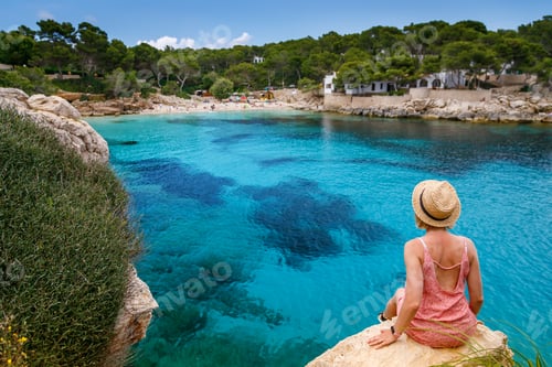 Preview: A woman leisurely sitting on a cliff, looking out at Cala Gat beach in Mallorca