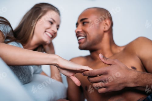 Preview: smiling multicultural couple holding hands in bed