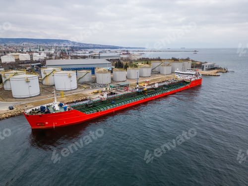 Preview: A red tanker ship docked at an industrial port with large storage tanks, surrounded by cityscape and