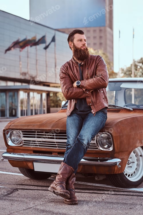 Preview: Bearded male dressed in brown leather jacket and boots with tuned retro car