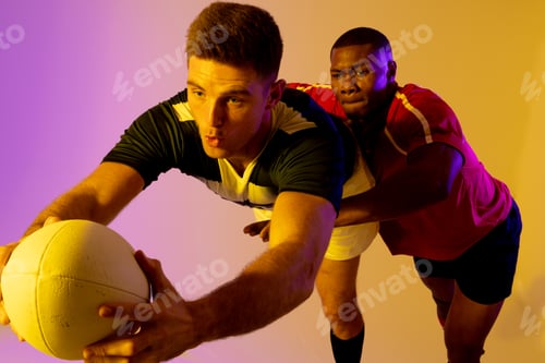 Preview: Diverse male rugby players with rugby ball playing over pink lighting