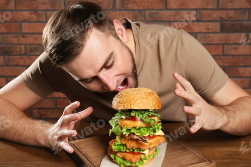 Preview: Young hungry man eating huge burger at table