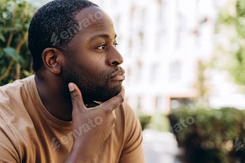 Preview: Calm young African American guy sitting in the city holding his hand on the chin