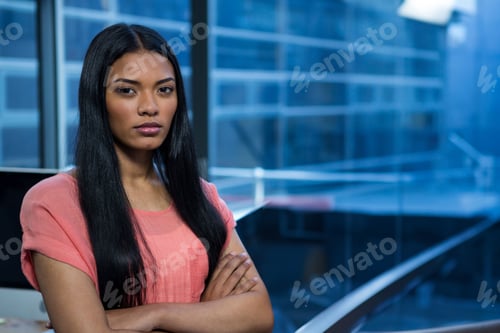Preview: African American woman wearing coral blouse standing with arms crossed in office beside monitor