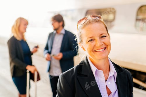 Preview: Smiling businesswoman at the train station with colleagues in the background
