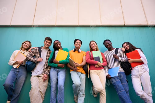 Preview: Low angle portrait of young multiracial students leaning against blue wall looking smiling at camera