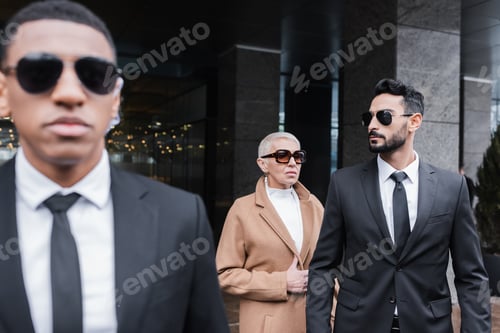 Preview: multiethnic security men in sunglasses protecting mature business lady near hotel building