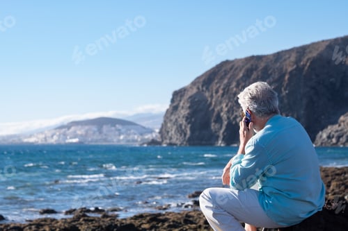 Preview: Senior man sitting at sunset light on a rocky beach face the ocean using mobile phone
