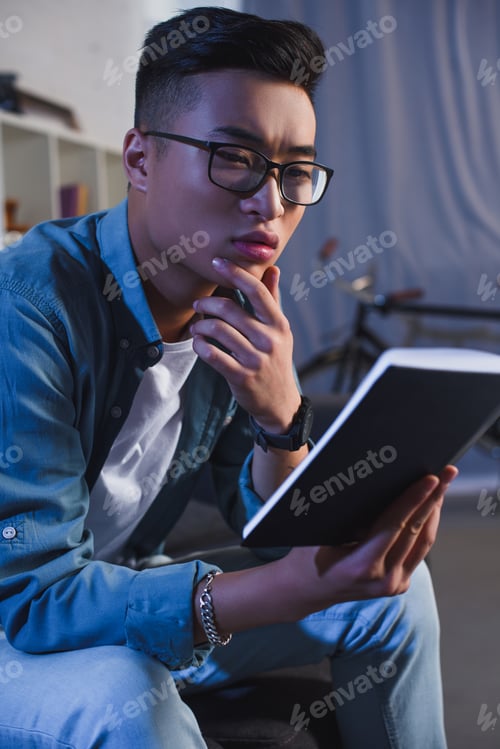 Preview: focused young asian man in eyeglasses reading book at home