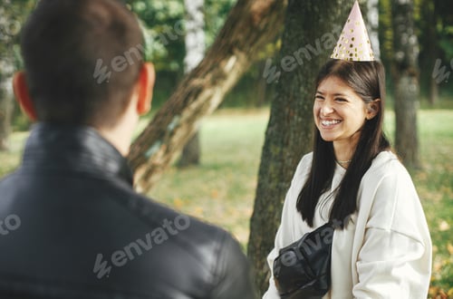 Preview: Stylish young happy woman in party hat celebrating birthday with family and friends at picnic
