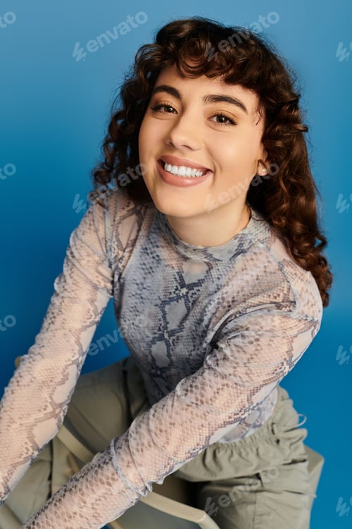 Preview: A young stylish woman expresses joy with her radiant smile in a trendy outfit, set against blue