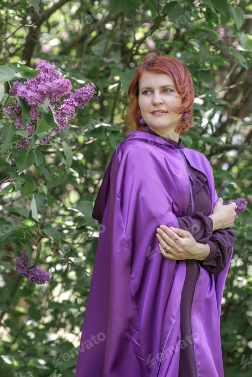 Preview: A beautiful young woman with red hair, against a background of blooming lilacs, in a cloak and hood.