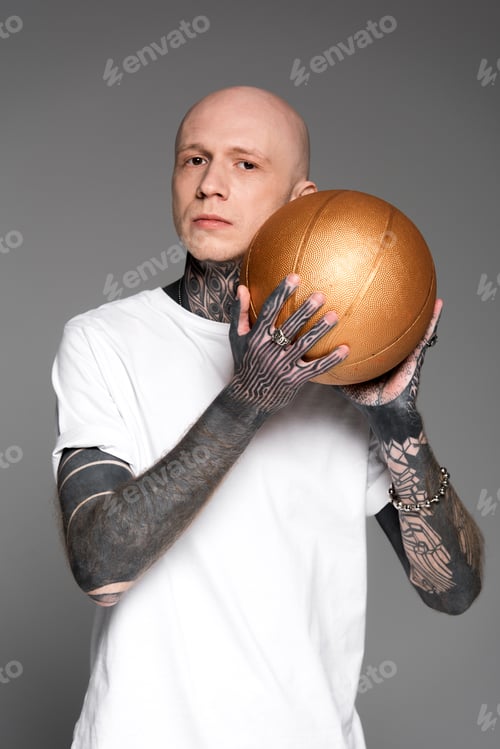 Preview: bald tattooed man in white t-shirt holding golden basketball ball and looking at camera isolated on