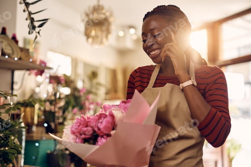 Preview: Happy black florist talking on cell phone while holding bouquet of flowers at flower shop.