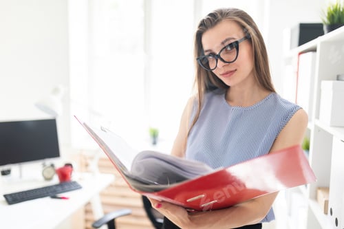 Preview: Beautiful young girl in an office stands near a rack and scrolls through a folder with documents.
