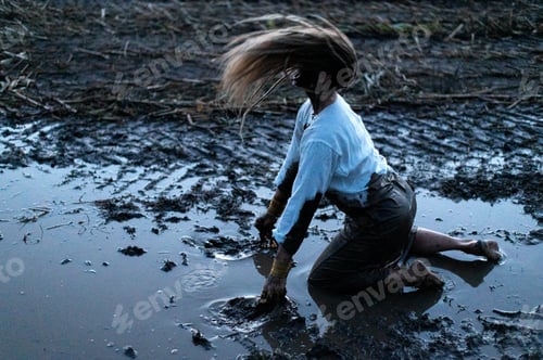 Preview: Dramatic portrait of a young woman kneeling in the black mud on field, cry of despair