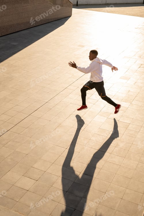 Preview: Young active sportsman in hoodie, sneakers and pants running down tiled pavement