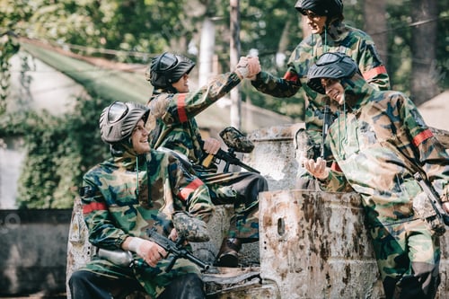 Preview: happy paintball team in camouflage celebrating victory and shaking hands on broken car outdoors