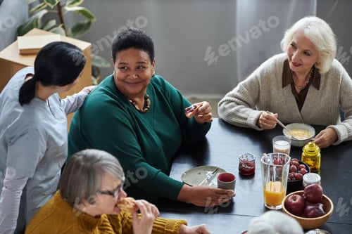 Preview: Seniors at breakfast in retirement home with young nurse assisting