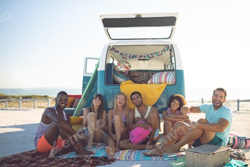 Preview: Happy group of diverse friends looking at camera while sitting near camper van at beach