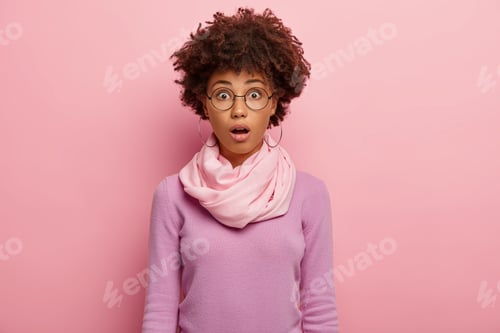 Preview: Emotional stupefied woman with curly Afro hairstyle, keeps mouth opened, wears round spectacles, cas
